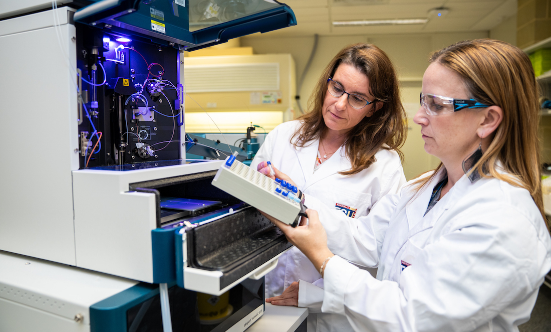 Professor Michelle Colgrave stand in front of laboratory equipment with associate