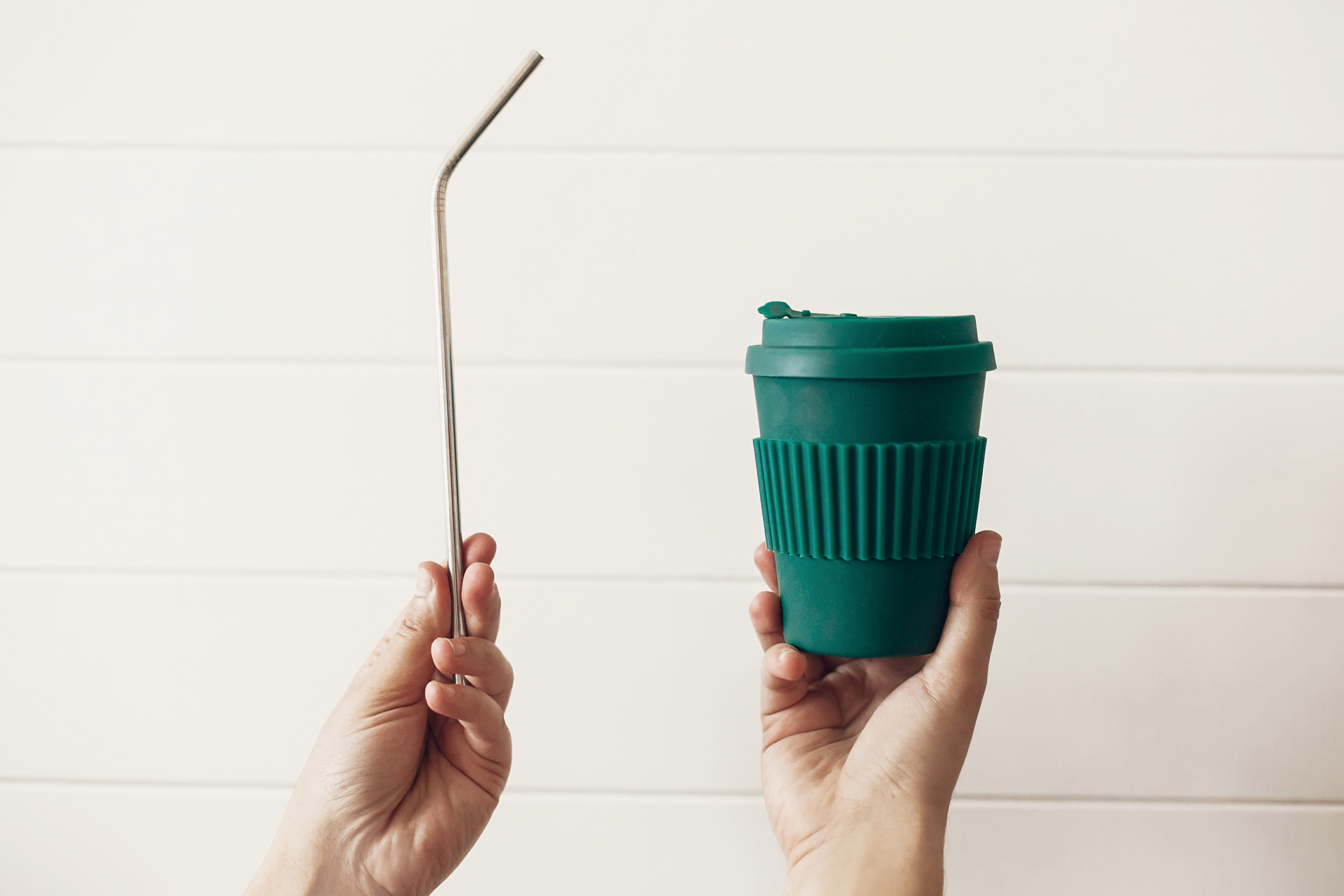 Hands holding stylish reusable eco coffee cup and steel straw on white wooden background