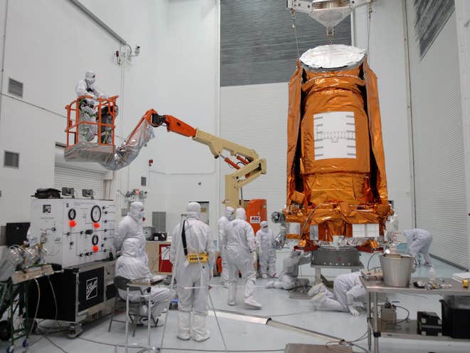 The large orange spacecraft on a stand for fuelling while workers in white suits stand beneath it