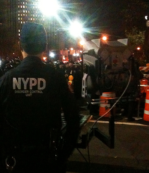 A police officer wearing a NYPD jackets stands next to a large speaker at a protest