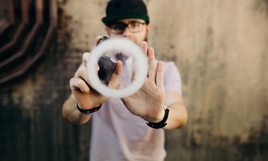 A young man wearing a cap holds out his hands and an e-cigarette to look like he's pushing a smoke ring forward