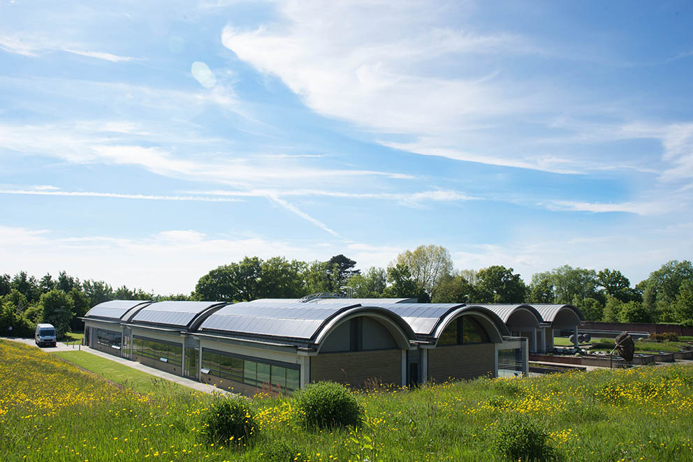 A series of long buildings stand in a field under a blue sky - it is the UK Native Seed Collection dedicated to Sir David Attenborough