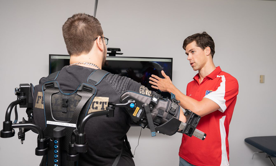 A researcher wearing a red t-shirt talks a participant through how to use the MindPod exoskeleton, which looks like a backpack vest that conects to the wearers' arms