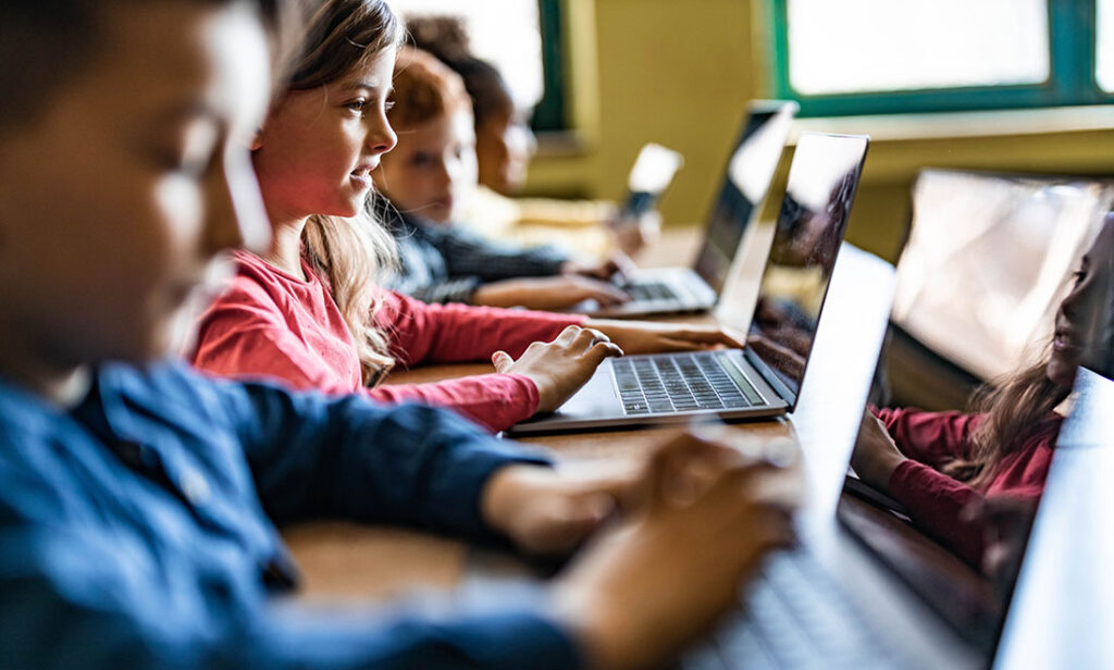 Happy schoolgirl and her classmates e-learning over laptops during a class in the classroom.