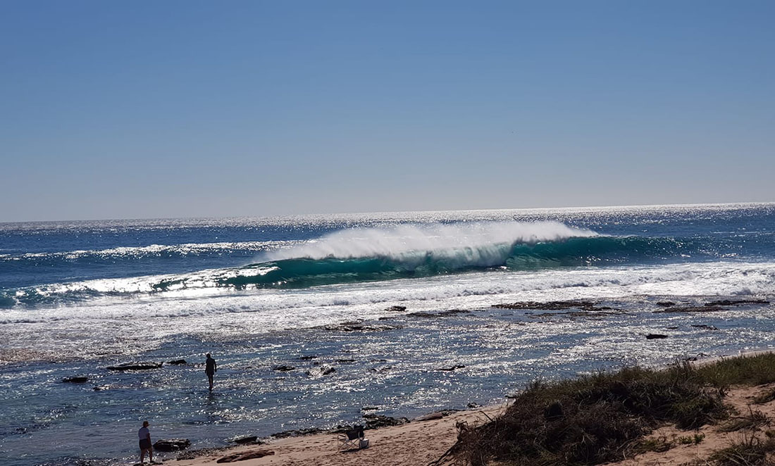 A wave crashes onto Kalbarri Beach as people swim in the shallow waters