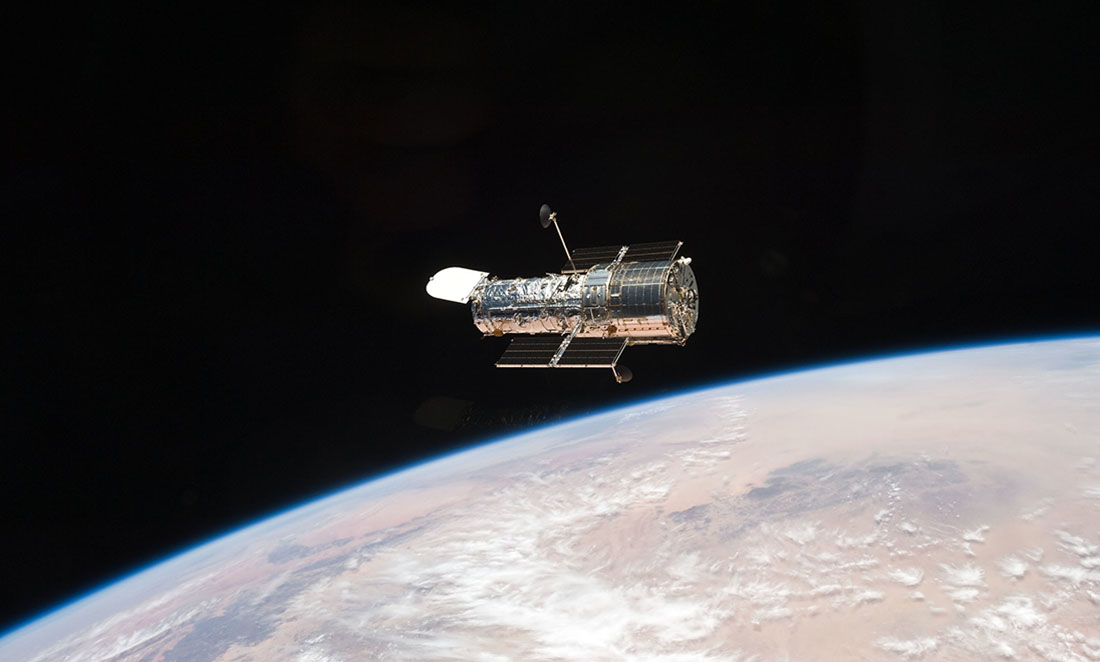 The large Hubble telescope shown against the darkness of space, floating above the surface of the Earth.