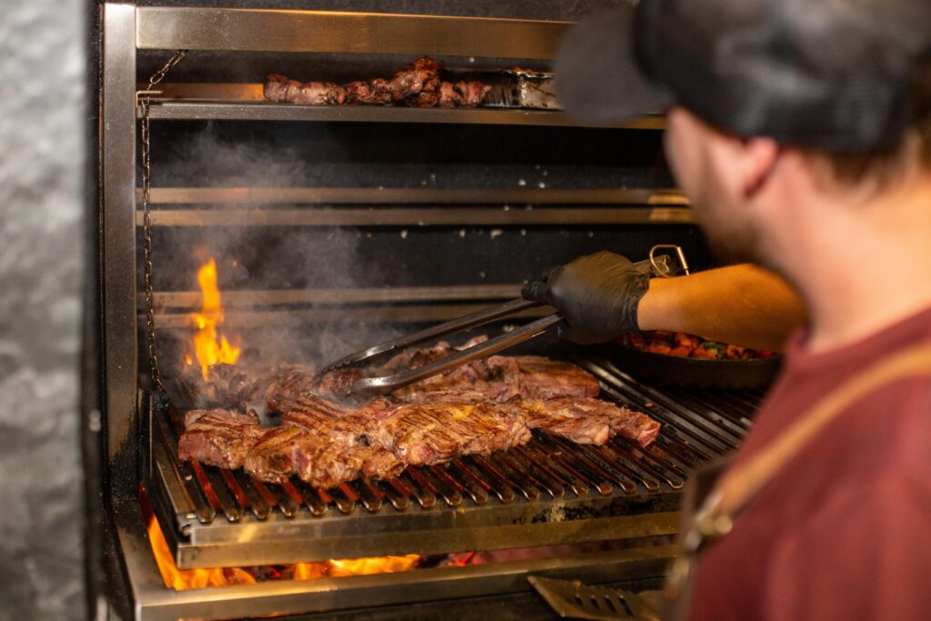 A photo looking over a cook's shoulder to a flame grill where meat is cooking