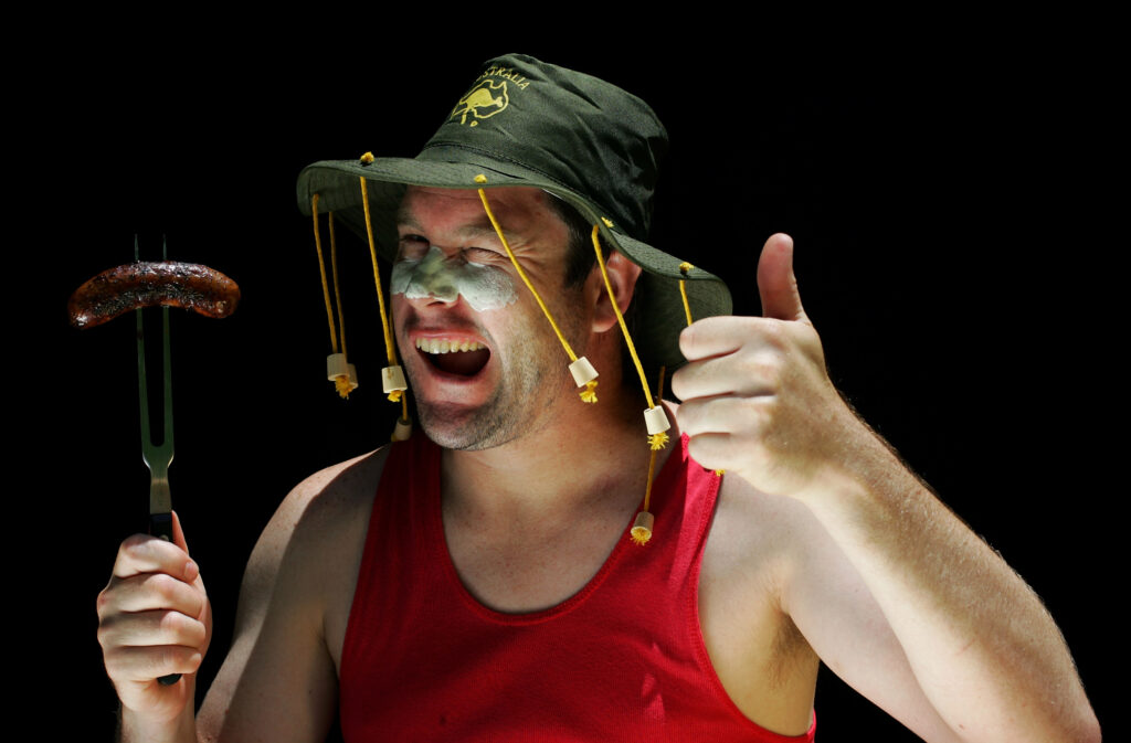 An Aussie man in a cork hat winks at the camera holding up a sausage on a fork in one hand and giving the thumbs up gesture with the other Credit:	Kolbz